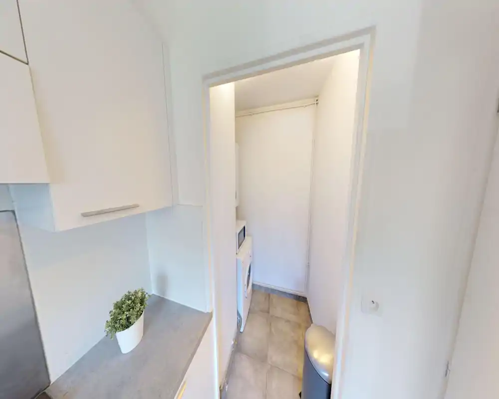 Angolo cucina compatto con pensili bianchi, piano grigio e una piccola pianta in vaso; dalla porta si intravede un vano tecnico con lavatrice e microonde.