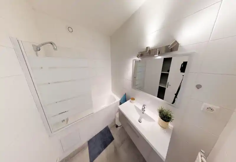 Salle de bain moderne et compacte avec baignoire-douche, murs carrelés blancs, meuble vasque avec miroir et éclairage clair et net.