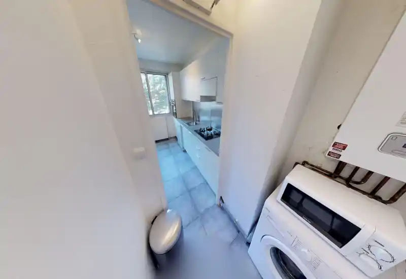 Cuisine en enfilade compacte avec placards blancs, crédence en inox et plaque à gaz ; une fenêtre apporte une belle lumière naturelle rendant l'espace fonctionnel.