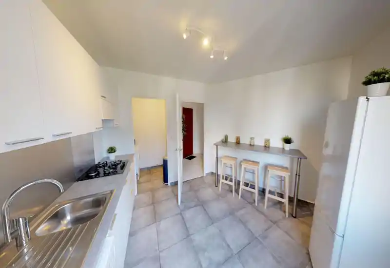 Cuisine moderne et lumineuse avec un long plan de travail, évier et plaque à gaz, ainsi qu’un bar avec trois tabourets — style épuré et agencement pratique.