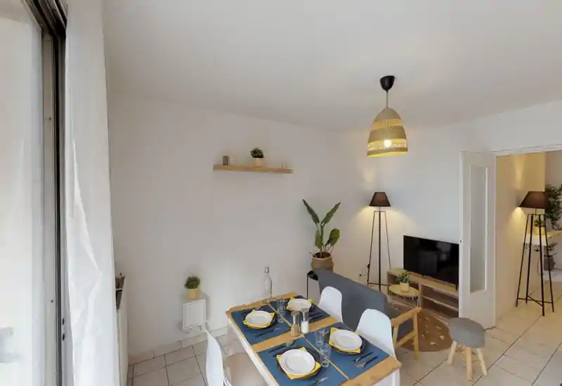 Bright, stylish living and dining area with a small sofa facing a TV, a wooden dining table set for four, warm lamps and green plants creating a cozy atmosphere.