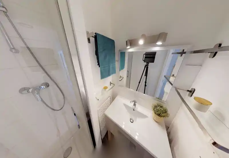 Salle de bain moderne et compacte avec douche vitrée, meuble vasque blanc et miroir, éclairée par un luminaire mural et une serviette turquoise — propre et bien éclairée.