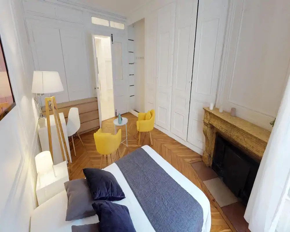 Bright, stylish bedroom with a large bed in the foreground, wooden herringbone floors and two cheerful yellow armchairs by a small table near a decorative fireplace.
