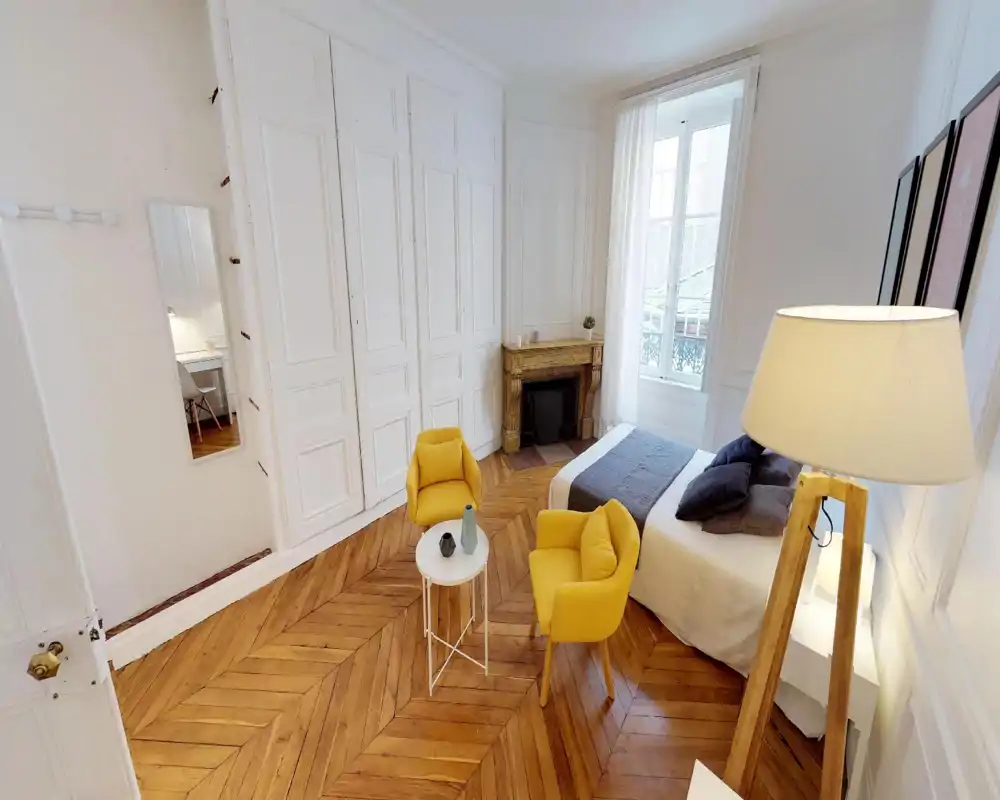 Bright, stylish bedroom with a comfortable double bed, decorative fireplace, and two yellow accent armchairs on warm herringbone parquet. Large window provides generous natural light; modern textiles and framed wall art create a welcoming atmosphere.
