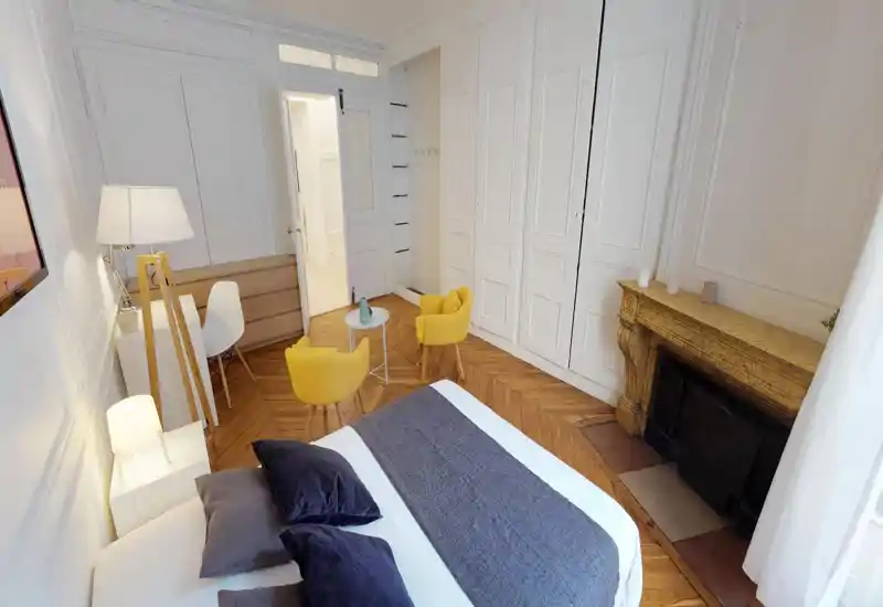 Bright, stylish bedroom with a large bed in the foreground, wooden herringbone floors and two cheerful yellow armchairs by a small table near a decorative fireplace.