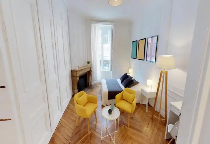 Bright, stylish bedroom with a double bed, bedside tables and cozy seating. Large window and floor lamp provide generous natural and artificial light; warm wooden herringbone floor and decorative fireplace add character.