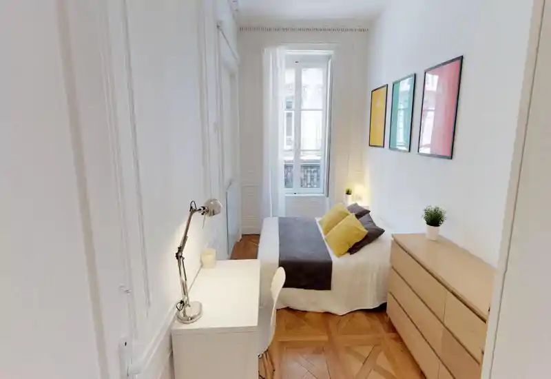 Bright, stylish bedroom with a double bed, bedside lamps, decorative cushions, framed wall art and a wooden chest of drawers; parquet flooring and a large window provide warm natural light.