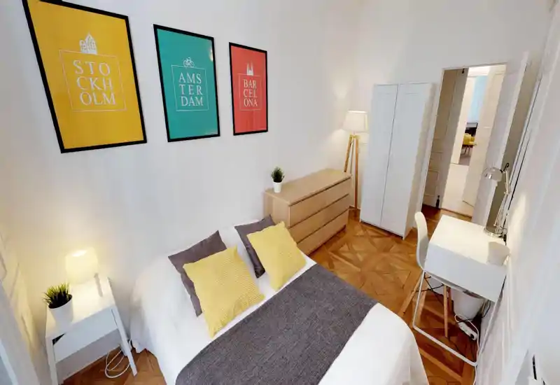 Bright, stylish bedroom with a double bed dressed in white linen, yellow accent pillows and a grey runner. The room features wooden parquet, a dresser, wardrobe and a small desk — cozy, well-lit and neatly staged for rentals.