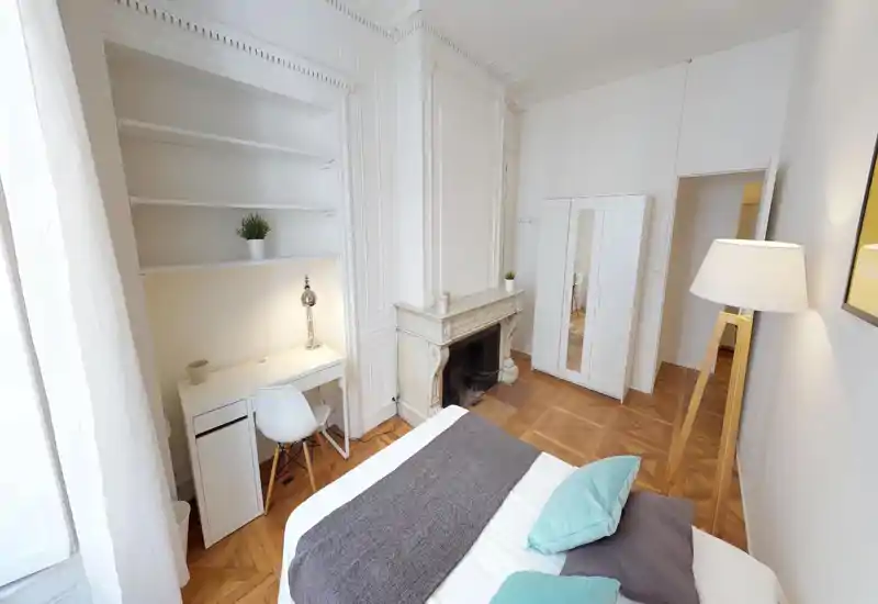 Bright, elegant bedroom with a visible double bed, decorative pillows and throw, white built-in shelving, a small desk and a classic fireplace. The room features warm parquet flooring and a tripod floor lamp for ambient light.