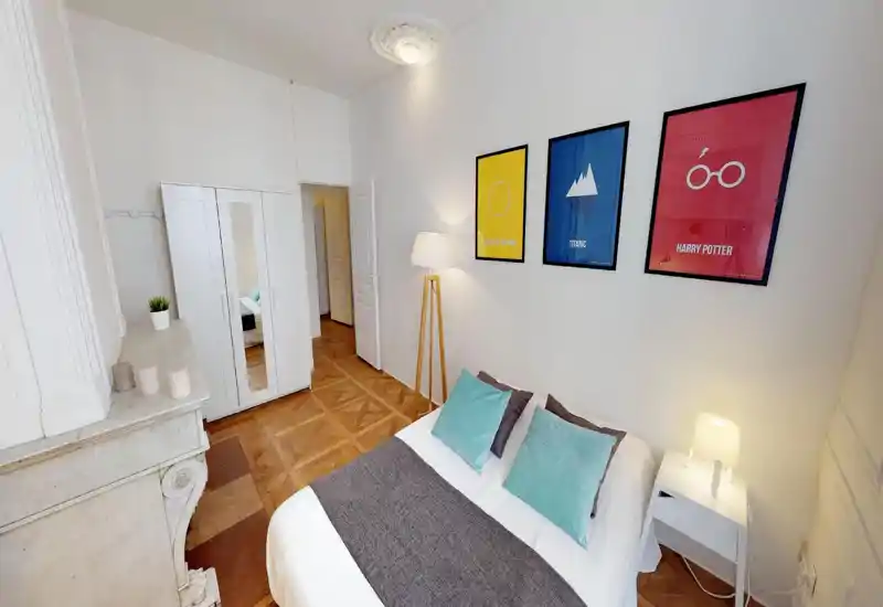 Bright, stylish bedroom with a double bed dressed in white linens, turquoise accent pillows and a gray runner; framed colorful posters and a standing lamp add personality.