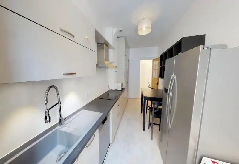 Modern galley kitchen with sleek white cabinets, dark stone countertop, integrated induction hob and stainless-steel fridge; bright, well-lit space with a small dining table and stools.