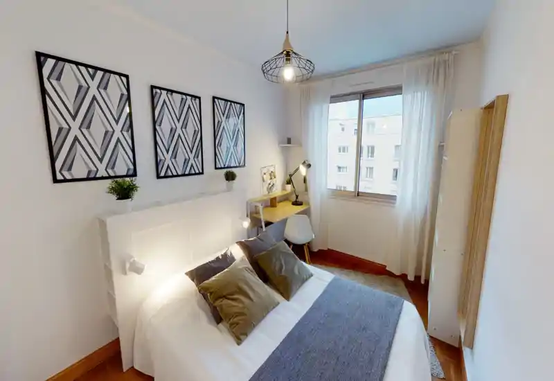 Bright, well-staged bedroom with a full bed in the foreground, decorative cushions, bedside lamps and a small desk by the window offering natural light.