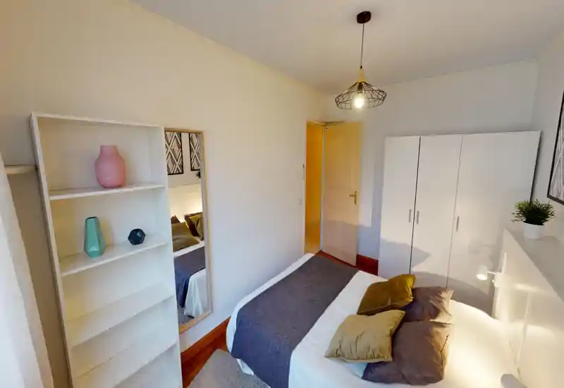 Bright, well-styled bedroom with a double bed dressed in neutral linens and decorative cushions, a white wardrobe and shelving, and a full-length mirror adding depth.