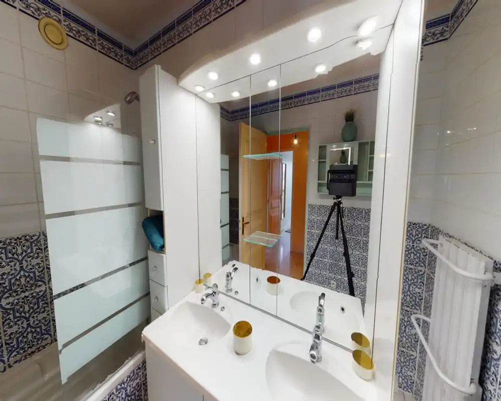 Bright, tiled bathroom with a double sink vanity, large mirror and shower/tub with frosted glass screen. Decorative blue patterned tiles and good overhead lighting create a clean, well-lit space.