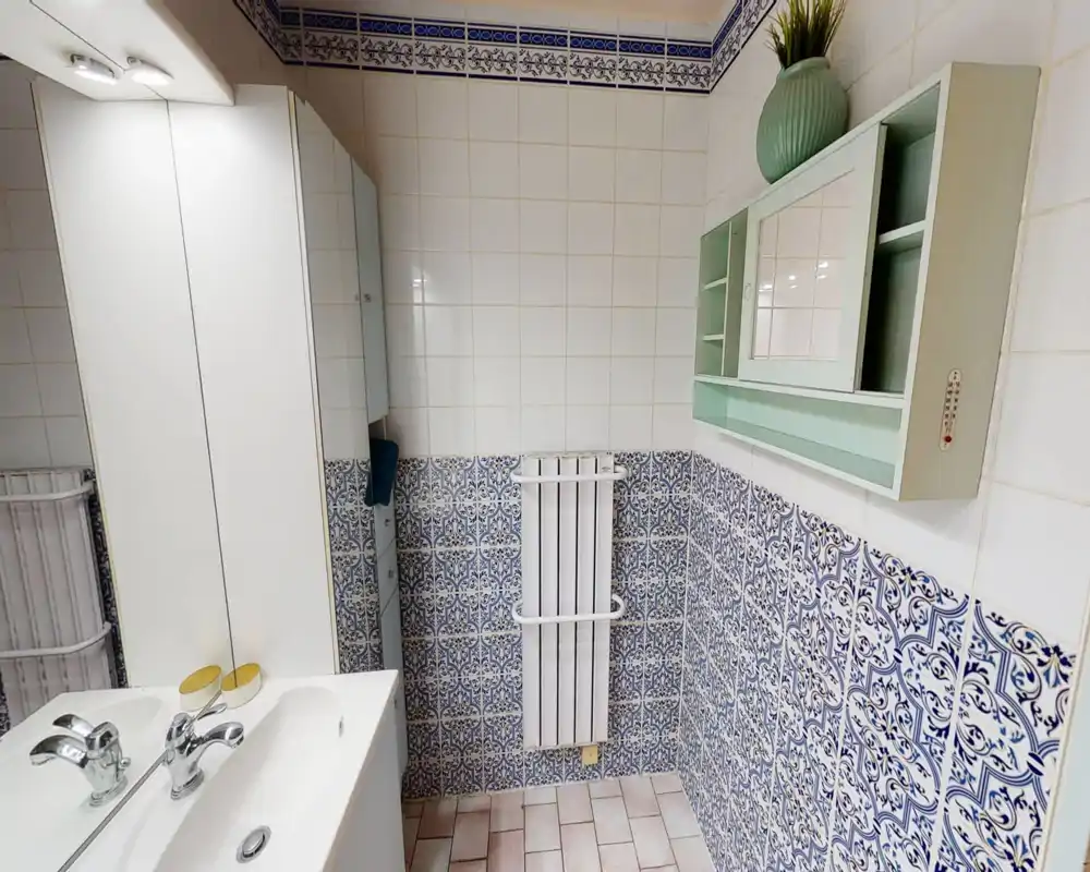 Bright, tiled bathroom with a double sink, mirrored cabinet and decorative blue patterned lower tiles. The space appears clean and well lit with a small radiator and wall storage.