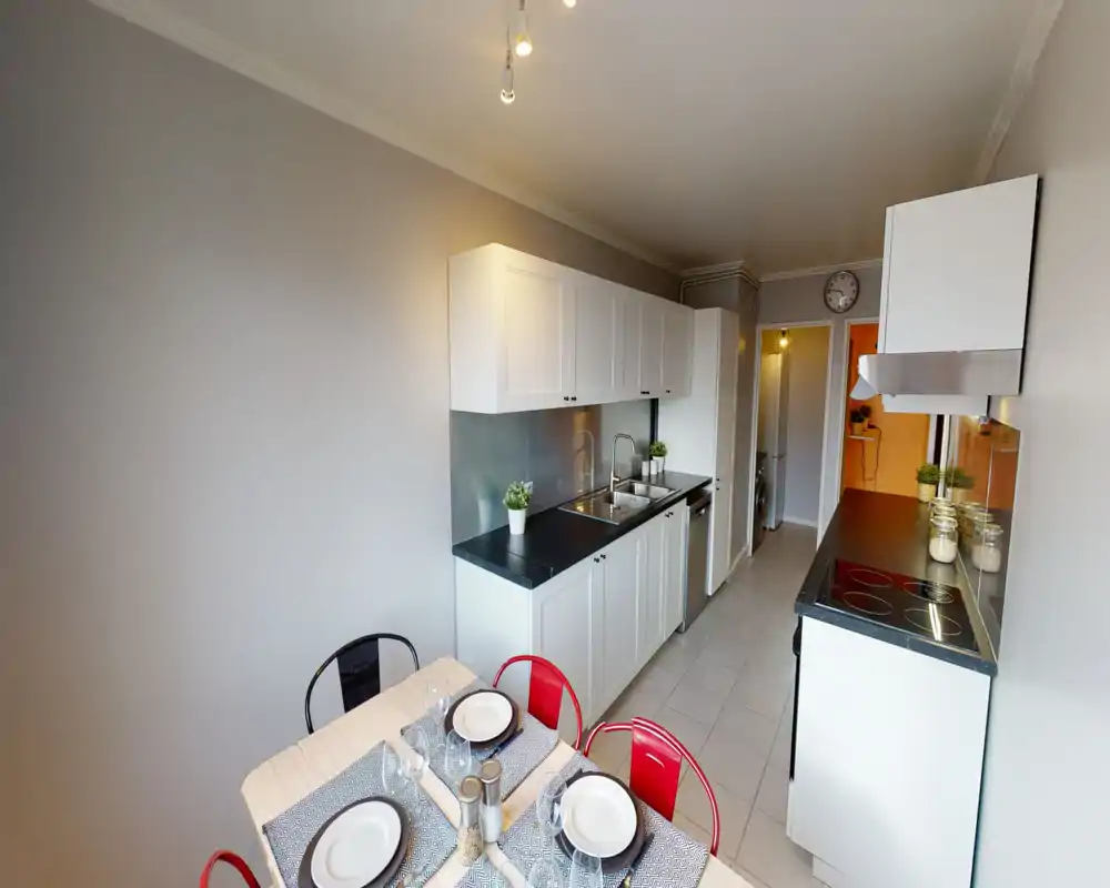 Bright, modern galley kitchen with white cabinets, black countertops, built-in sink and hob. A small dining table set for four and decorative plants create a welcoming, well-staged space.