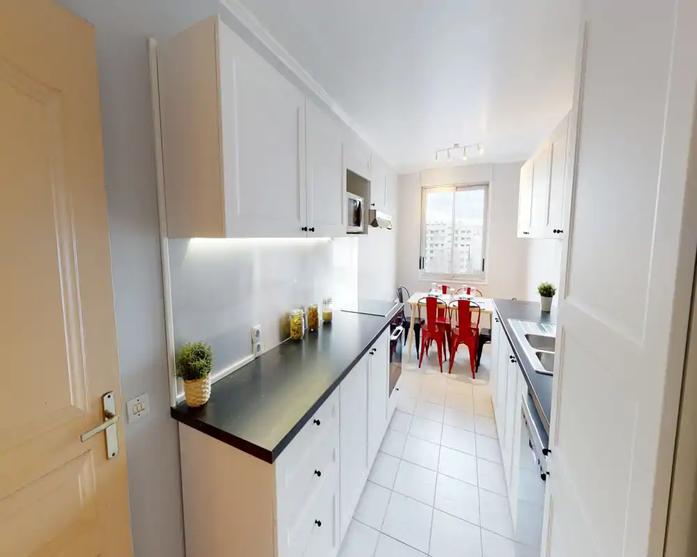 Bright modern kitchen with white cabinets, black countertops, built-in appliances and a small dining area by the window — neat decorative jars and plants add warmth.