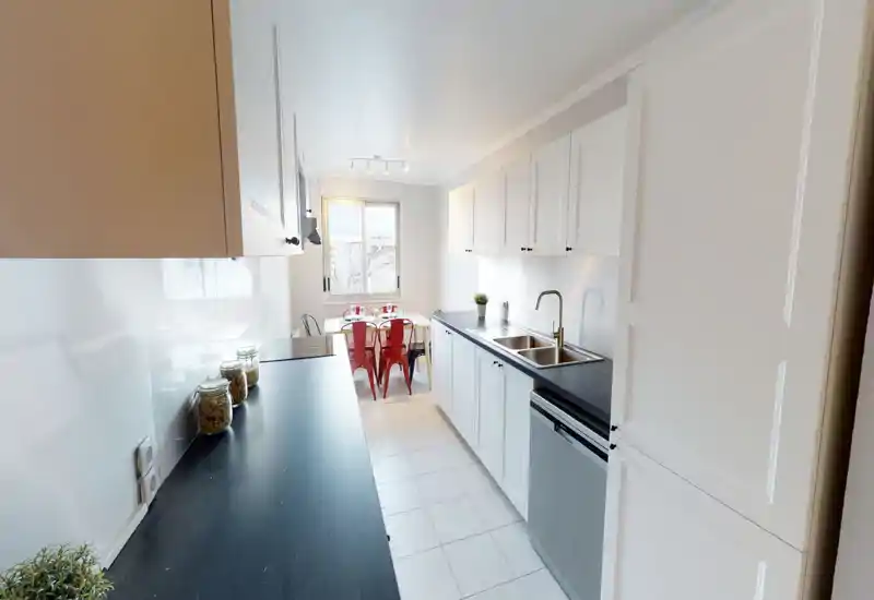 Bright, modern galley kitchen with white cabinets, dark countertops, a stainless sink and a small dining nook with red chairs by the window.