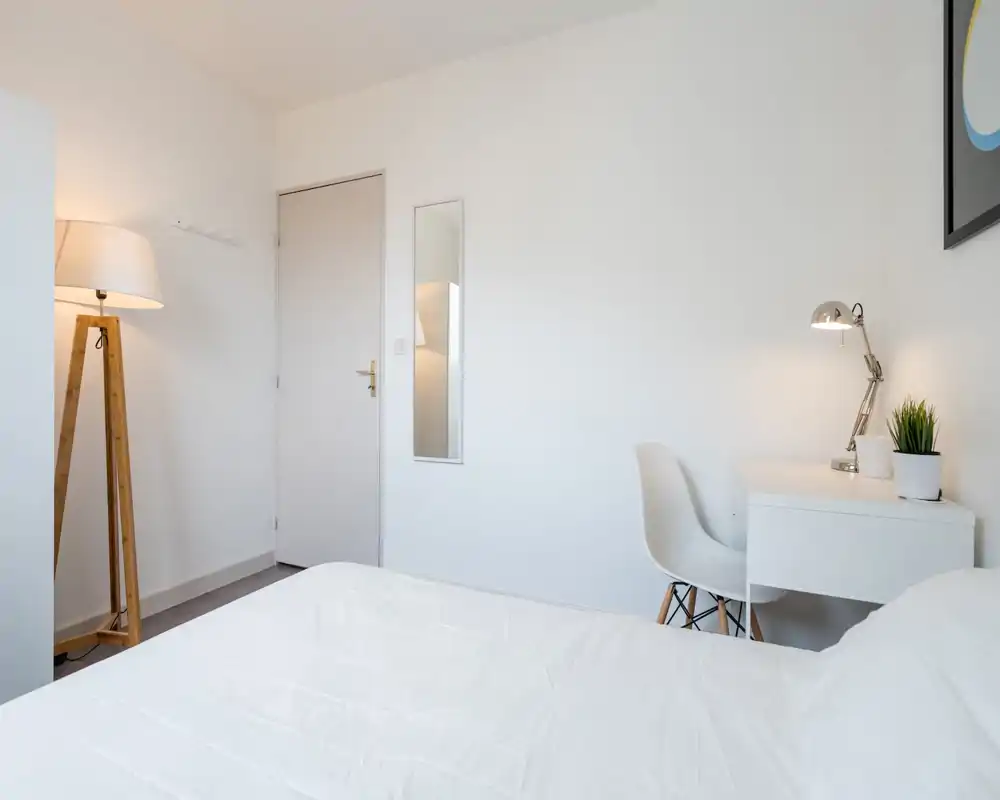 Chambre minimaliste et lumineuse avec lit blanc au premier plan, lampe sur trépied en bois et petit bureau blanc avec chaise et lampe — propre, moderne et aérée.