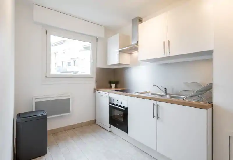 Cuisine compacte et moderne, lumineuse, avec placards blancs, four et lave-vaisselle intégrés, évier double et plan de travail en bois — propre et bien éclairée par une grande fenêtre.