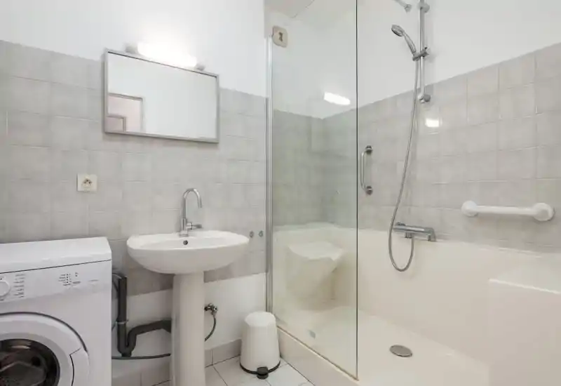 Salle de bains lumineuse et moderne avec une douche séparée par une paroi en verre, lavabo sur colonne et lave-linge. Carrelage gris neutre et robinetterie chromée pour un rendu propre et fonctionnel.