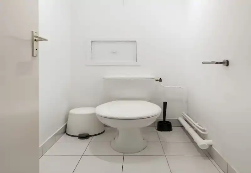 Petite salle d’eau blanche avec WC standard, brosse et petite poubelle ; agencement lumineux, minimaliste et propre, parfait pour un WC invités.