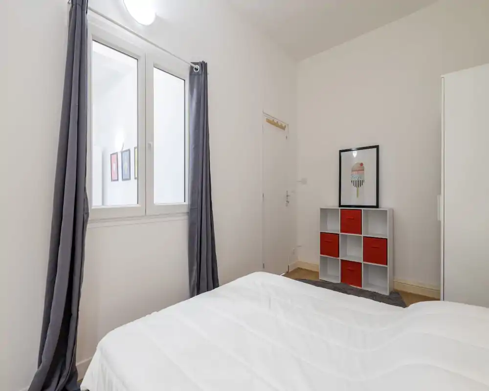 Camera da letto luminosa e minimalista con un letto bianco rifatto in primo piano, tende grigie e un piccolo mobile a cubi con contenitori rossi — stile pulito e moderno.