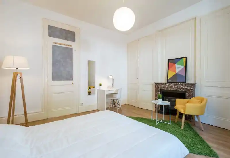 Camera da letto luminosa e dallo stile moderno con un ampio letto bianco in primo piano, una poltrona gialla accogliente, tappeto verde e una piccola scrivania vicino alla parete. Toni neutri, soffitti alti e illuminazione curata creano un’atmosfera ariosa e invitante, perfetta per annunci.