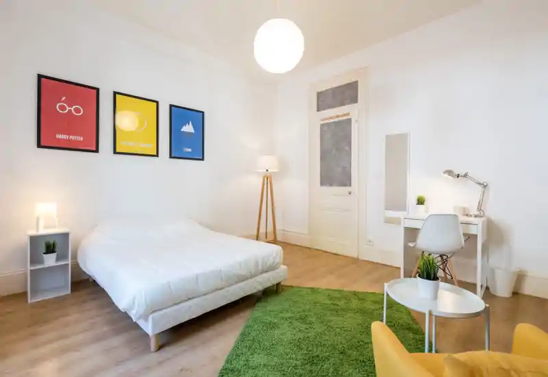 Camera da letto moderna e luminosa con letto matrimoniale bianco, comodino e scrivania. Arredamento minimal, stampe colorate e un tappeto verde che donano calore.