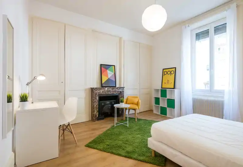 Camera da letto moderna e luminosa con letto bianco, tappeto verde e scrivania dal design minimale. La grande finestra garantisce luce naturale e dettagli di arredo (poltrona gialla, stampe incorniciate) la rendono accogliente e appetibile per annunci.