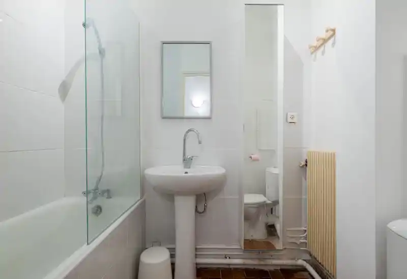 Clean, compact bathroom with a bathtub and shower screen, pedestal sink, wall mirror and a toilet visible in a tall mirror. Neutral white tiles and simple fixtures offer a functional, well-lit space.
