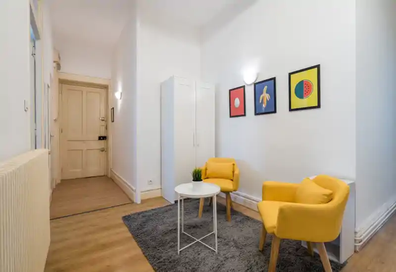 Bright, tidy entrance area with wooden flooring, two yellow accent chairs, a small white table and a wardrobe — well-lit and welcoming for guests.