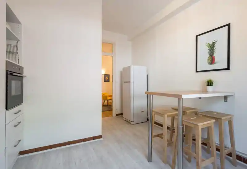 Compact kitchen area with a small breakfast bar, four wooden stools and a refrigerator; clean, minimalist decor with a framed print and a potted plant.