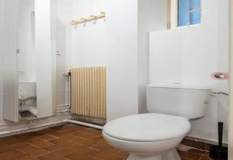 Small, functional bathroom with a white toilet, wall-mounted radiator and a mirrored cabinet. The space is simply finished with terracotta floor tiles and bright white walls.