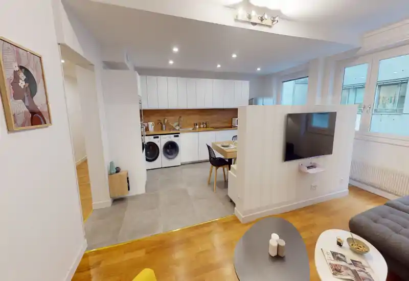 Espace de vie moderne et lumineux en open space avec une cuisine compacte. On distingue un canapé confortable, une table basse et une télévision murale séparant le salon de la cuisine/salle à manger.