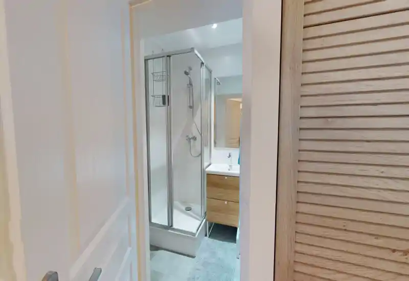 Salle de bains compacte et moderne avec cabine de douche d’angle, robinetterie murale et petit meuble vasque avec miroir — lumineux, propre et bien agencé.