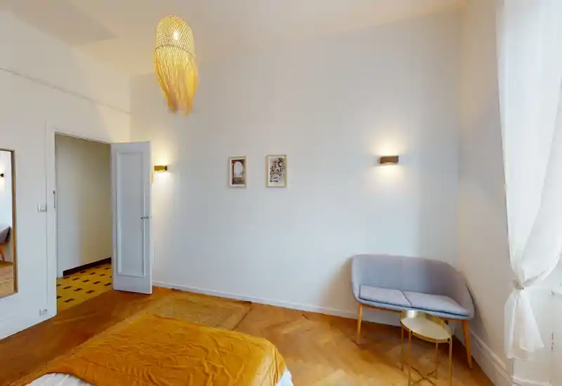 Camera da letto luminosa e di stile con pavimento in parquet caldo, copriletto color senape, una poltroncina vicino alla finestra e luci da parete eleganti — confortevole e ben illuminata.