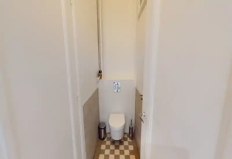 Toilettes compactes et lumineuses avec plaque de chasse murale, petite poubelle et brosse. Carrelage neutre et sol en damier pour un look propre et contemporain.