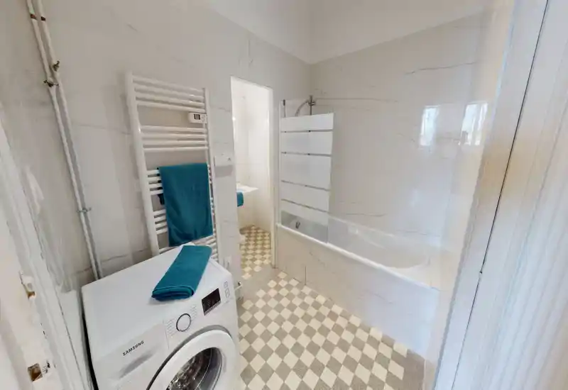 Salle de bains moderne et lumineuse avec baignoire/douche, carrelage à motif, radiateur porte-serviettes mural et machine à laver frontal.