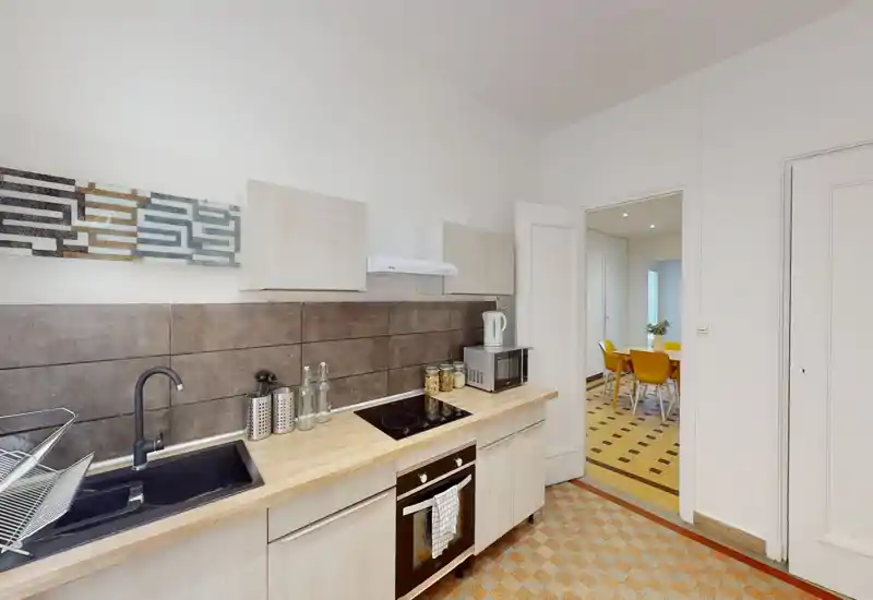 Cuisine moderne et lumineuse avec plan de travail en bois, four encastré et plaque à induction, et évier noir. Bocaux décoratifs et ustensiles apportent une touche conviviale ; la porte ouvre sur une salle à manger avec des chaises jaunes.