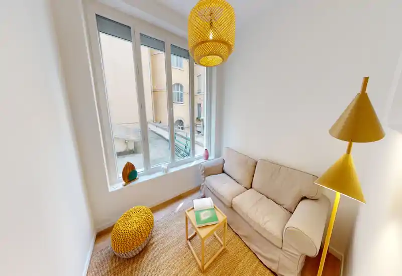 Soggiorno piccolo, luminoso e accogliente con divano neutro per due persone, ampia finestra che fornisce luce naturale e tocchi caldi gialli nella lampada e nel paralume. Tappeto intrecciato e pouf completano l'allestimento perfetto per un appartamento compatto.