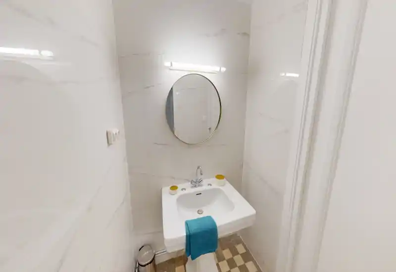 Salle de bains compacte et lumineuse avec lavabo sur colonne, miroir rond et carrelage moderne — propre et bien éclairée, adaptée comme toilette d'appoint.