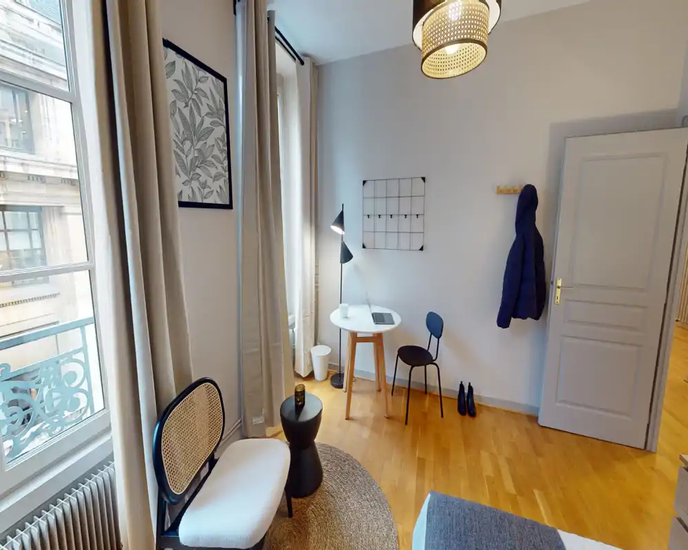 Coin chambre lumineux et élégant avec un petit espace d'assise, un bureau et une vue partielle du lit. Grandes fenêtres et parquet chaleureux pour une ambiance accueillante.