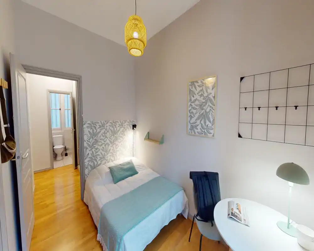 Chambre simple lumineuse et confortable avec tête de lit décorative, sol en bois et éclairage de chevet. La pièce comprend un petit espace de travail avec table ronde et chaise, et une salle de bain attenante visible par la porte ouverte.