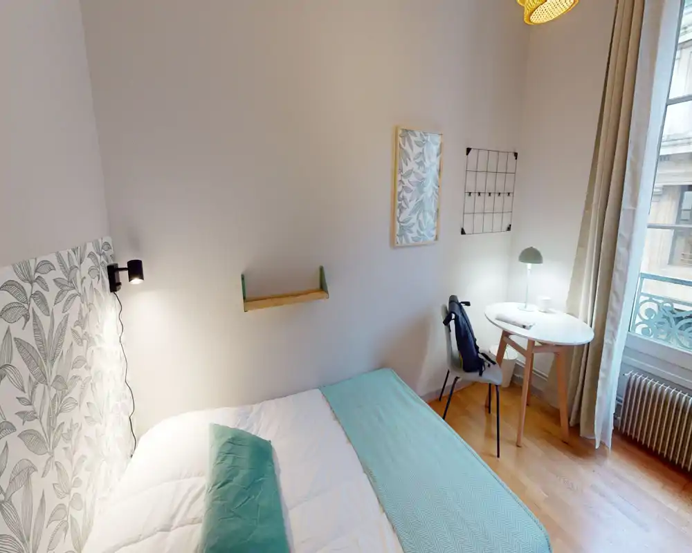 Chambre lumineuse et chaleureuse avec lit double, tête de lit décorative et petite table ronde près de la fenêtre. La grande fenêtre apporte une belle lumière naturelle; accents turquoise et parquet donnent une ambiance paisible.