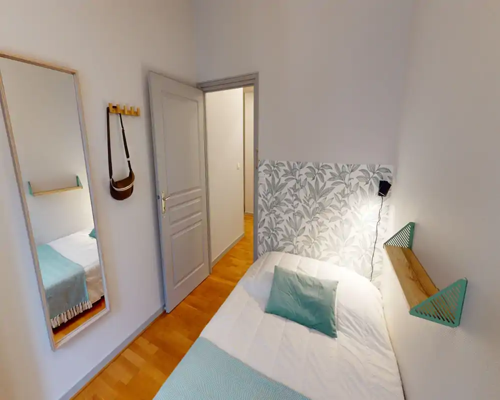 Chambre compacte et lumineuse avec un lit simple confortable, une tête de lit partielle à motif et des accents bleu-vert. Un miroir sur pied et des crochets muraux offrent du rangement pratique; le sol en bois apporte une atmosphère chaleureuse.