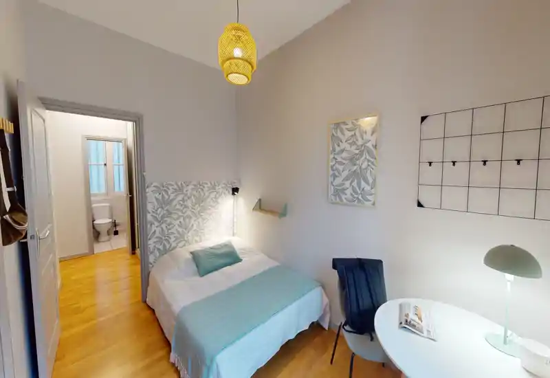 Bright, cozy single bedroom with a decorative headboard, wooden floor and bedside lighting. The room includes a small workspace with a round table and chair and an ensuite bathroom visible through the open door.