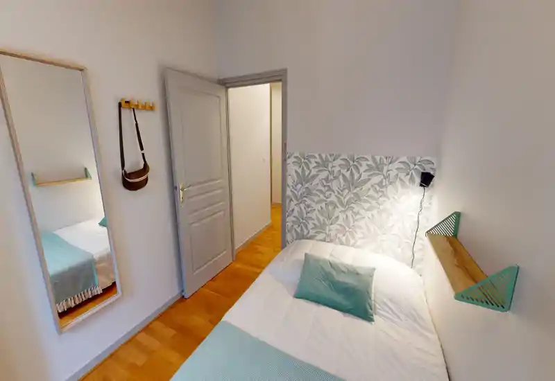 Small, bright bedroom with a comfortable single bed, patterned half-headboard and soft teal accents. A full-length mirror and wall hooks add functionality; warm wooden floors complete the cozy look.