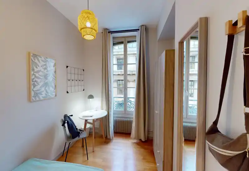 Bright, neatly staged bedroom with a visible bed corner, a small desk by the window and warm wooden floors—ideal for a single occupant or short stays.