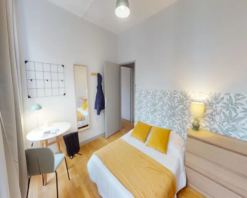 Bright, neatly staged single bedroom with a mustard-accented bed, bedside chest, small work table and soft natural light — ideal for a cozy rental listing.